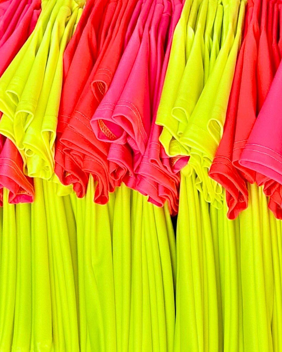 Product image of bright swimwear, short-sleeve neon rash guards, handing with sleeves facing the camera. Neon pink rash guard, neon red rash guard, neon yellow rash guard.